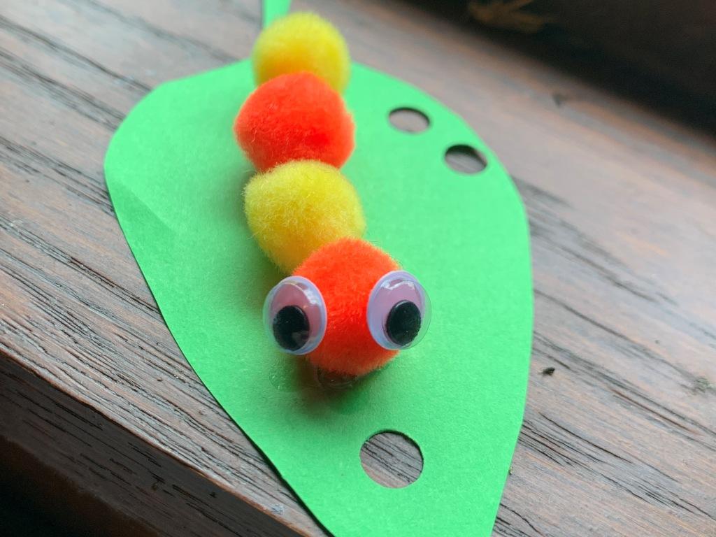 This photo of a finished craft, a caterpillar, made with fuzzy pom pom balls and wiggly eyes, on a leaf made of construction paper.